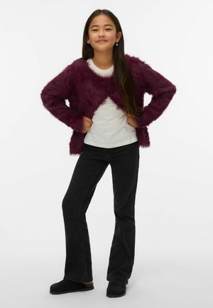 Mīksts bordo kardigans, balta krekls un melnas izplatītas džinsi; modele pozē, rokas uz gurniem; melnas slip-on kurpes noslēdz izskatu.