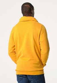 Pull jaune avec un col montant, fabriqué en tissu doux. Il présente des manches longues et une coupe décontractée, porté avec un jean bleu. Pas de graphismes visibles.