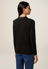 Schwarzes Langarmoberteil aus glatter Stoff, mit einem schlichten Design und geradem Schnitt. Kombiniert mit blauen Jeans, die eine klare Silhouette zeigt.