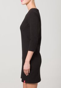 Musta ribbineulottu mekko, jossa on kolme neljänneksen hihaa, vartalonmyötäinen muoto ja sivuhalkio. Helmassa pieni punainen logo. Sileä koostumus.