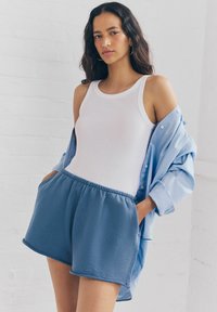 Canottiera bianca con spalline larghe, abbinata a pantaloni corti blu larghi con vita elastica e tasche, indossata con una camicia azzurra a bottoni.