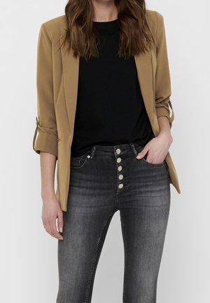 Vrouw draagt een beige blazer over een zwarte top met een high-waisted grijze spijkerbroek met vijf knopen aan de voorkant, hand in de zak.