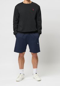 Svart crew-neck sweatshirt med röd logotyp, kombinerat med marinblå shorts. Modellen bär vita sneakers och vita strumpor som går till ankeln.