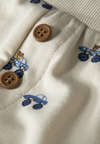 Světle béžová tkanina obsahuje vyšívané modré traktory a hnědé medvědy, uprostřed jsou dvě hnědé knoflíky. Je viditelná jemná struktura.