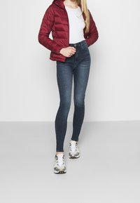 Burgundská zateplená bunda s kapucí, bílý přiléhavý top, tmavé skinny džíny a šedé tenisky se žlutými akcenty.