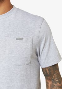 Camiseta de algodón gris con cuello redondo, mangas cortas y un bolsillo en el pecho izquierdo con un detalle de etiqueta plateada. Textura suave, diseño casual.