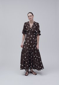 Mujer de pie con un vestido largo marrón oscuro con grandes lunares blancos, mangas abullonadas y sandalias negras de punta abierta sobre un fondo liso.