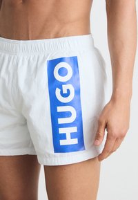 Shorts de bain blancs avec un grand logo bleu portant le mot "HUGO" sur le côté gauche. Le tissu semble léger et possède une taille élastique.