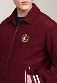 Giacca in corduroy bordeaux con zip, dotata di polsini a strisce bianche, tasche laterali e un logo ricamato sul petto.