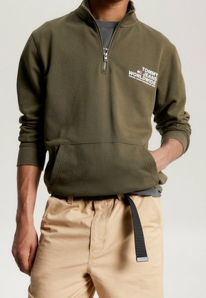 Olivgrüner Reißverschluss-Hoodie mit großem Fronttasche und weißem Textlogo. Er hat gerippte Bündchen und eine lockere Passform, über einem grauen Shirt getragen.