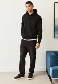 Felpa nera con cappuccio e design strutturato, dotata di coulisse, indossata sopra una maglietta bianca. Abbinata a jeans scuri e sneakers nere.