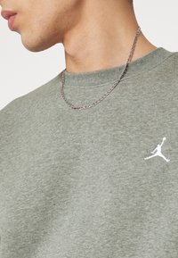 Primer plano de una persona que lleva un collar de cadena de plata y una camisa verde oliva con un pequeño logo de un jugador de baloncesto blanco.