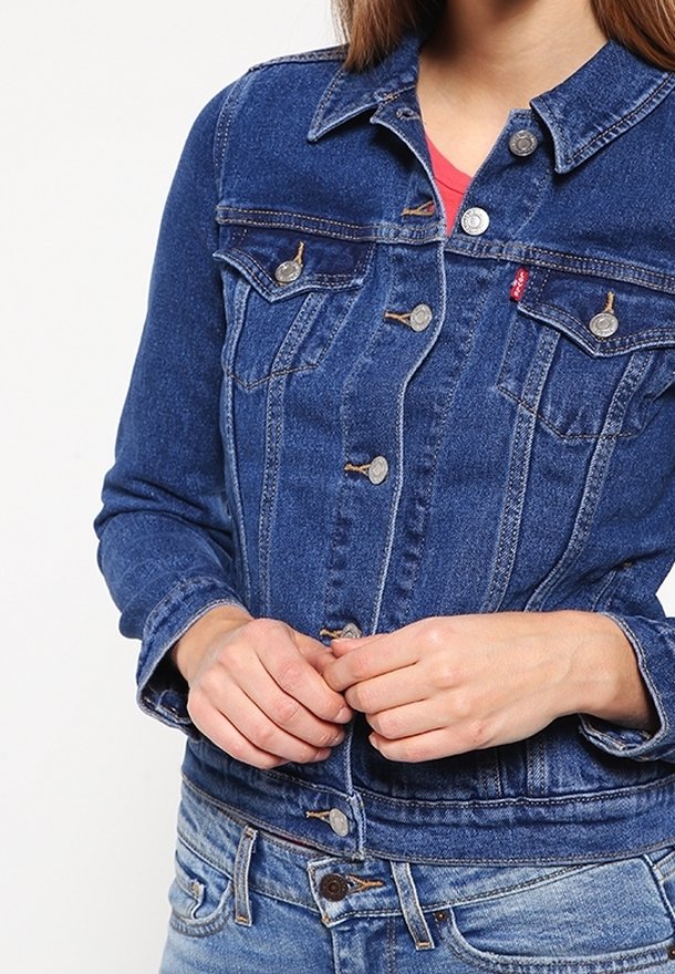 Nainen, joka pitää päällään sinistä farkkutakkia ja farkkuja, kiinnittää takin nappeja molemmilla käsillään.