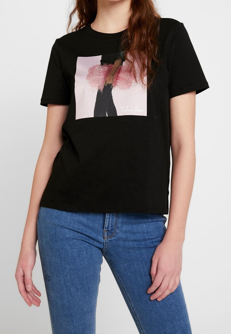 Jeune femme portant un t-shirt noir avec un motif tutu rose et un jean bleu, debout sur un fond blanc uni.