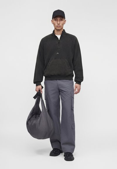 Άνδρας με μαύρο καπέλο, μαύρο φούτερ με φερμουάρ στο λαιμό, γκρι παντελόνι cargo, μαύρα παπούτσια, κρατώντας μεγάλη γκρι επενδεδυμένη τσάντα στο αριστερό χέρι.