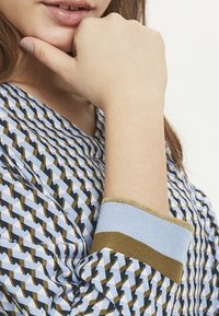 Kvinna som vilar hakan på handen, klädd i en mönstrad tröja i blått, brunt och vitt med randiga manschetter och delvis synliga läppar.