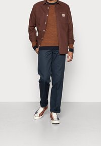 Man som bär en brun skjortjacka över en orange tröja, marinblå byxor och vita sneakers med bruna och marinblå detaljer som står på en grå golv.