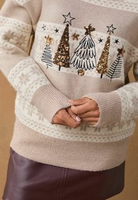 Maglione beige con tessuto testurizzato, caratterizzato da alberi ricamati e paillettes dorate. Maniche a costine e una fascia decorata completano il design.
