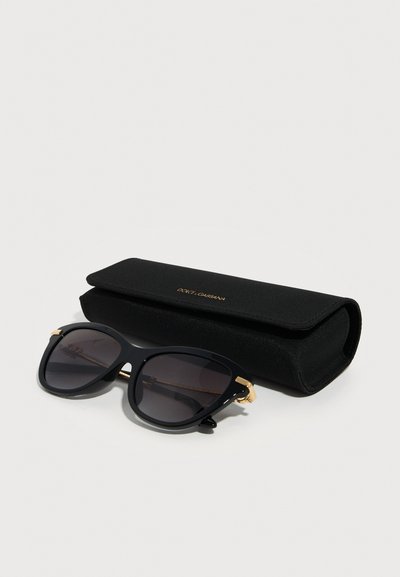 Μαύρα γυαλιά ηλίου με τη μορφή γάτας και χρυσές λεπτομέρειες δίπλα σε μαύρη ορθογώνια θήκη με ανάγλυφο χρυσό λογότυπο Dolce & Gabbana σε λευκή επιφάνεια.