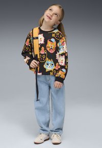 Sudadera negra con estampados de gatos coloridos, jeans azul claro holgados y zapatillas beige. Tirante de mochila amarilla cruzado sobre el hombro.