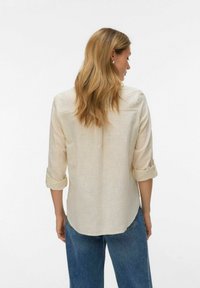 Femme aux longs cheveux blonds portant une chemise beige aux manches retroussées et un jean bleu, tournée de dos sur un fond blanc uni.