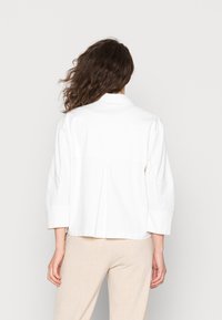 Chaqueta blanca de algodón con un corte relajado, mangas de 3/4 y un detalle de pliegue en la parte posterior. Diseño simple con una textura limpia y suave.