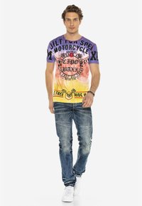 T-shirt graphique à manches courtes violet, orange et jaune avec des motifs sur le thème de la moto, associé à un jean bleu délavé et des baskets blanches.