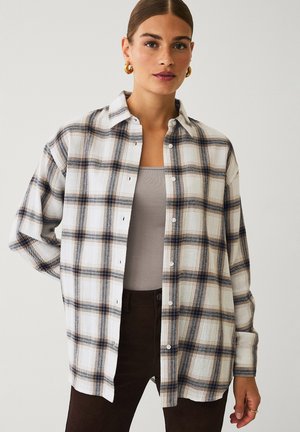 Kvinde iført en beige, brun og hvid ternet skjorte med knapper over en beige tanktop med mørkebrune bukser og gyldne hoops-øreringe.