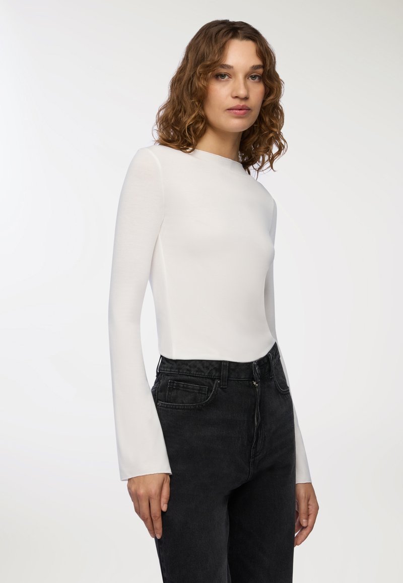 Maglietta bianca a maniche lunghe con collo alto, design aderente, abbinata a jeans neri a vita alta, realizzata in un tessuto leggero e morbido.