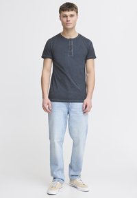Mørkeblå kortærmet t-shirt med rund halsudskæring, tre-knap lukning og op rullede ærmer, båret med lyseblå jeans og beige sneakers.