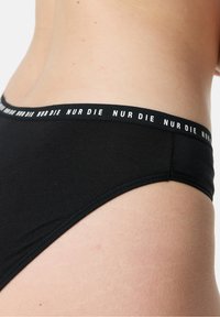 Black cotton bikini brief with a wide waistband featuring white "NUR DIE" text. Smooth texture, fitted design, and minimalistic style.
