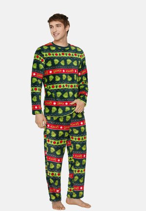 Młody mężczyzna uśmiechający się, ubrany w zielono-czerwone, wygodne świąteczne piżamy z motywem Grincha, stojący boso na jednolitym jasnym tle.