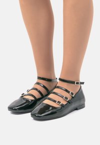Zapatos de charol negros con puntera puntiaguda, que cuentan con tres correas de hebilla a lo largo del pie y el tobillo. Suela plana y diseño minimalista.