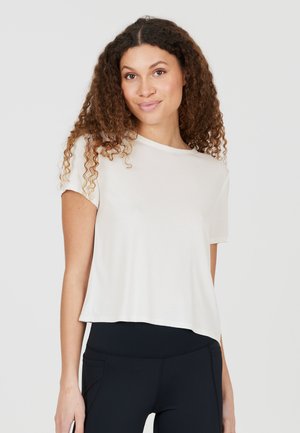 Femme aux cheveux bouclés portant un haut blanc ample à manches courtes et des leggings noirs taille haute sur un fond uni.