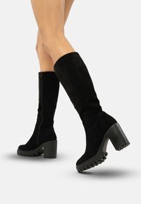 Botas altas de ante negro con suela de tacón grueso, que cuentan con un cierre de cremallera lateral y una suela de goma texturizada para mayor agarre.