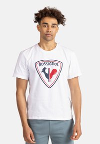 Camiseta de algodón blanca con un gran logo de escudo que presenta siluetas de gallos en negro y rojo y el texto "ROSSIGNOL" arriba. Diseño sencillo de cuello redondo.