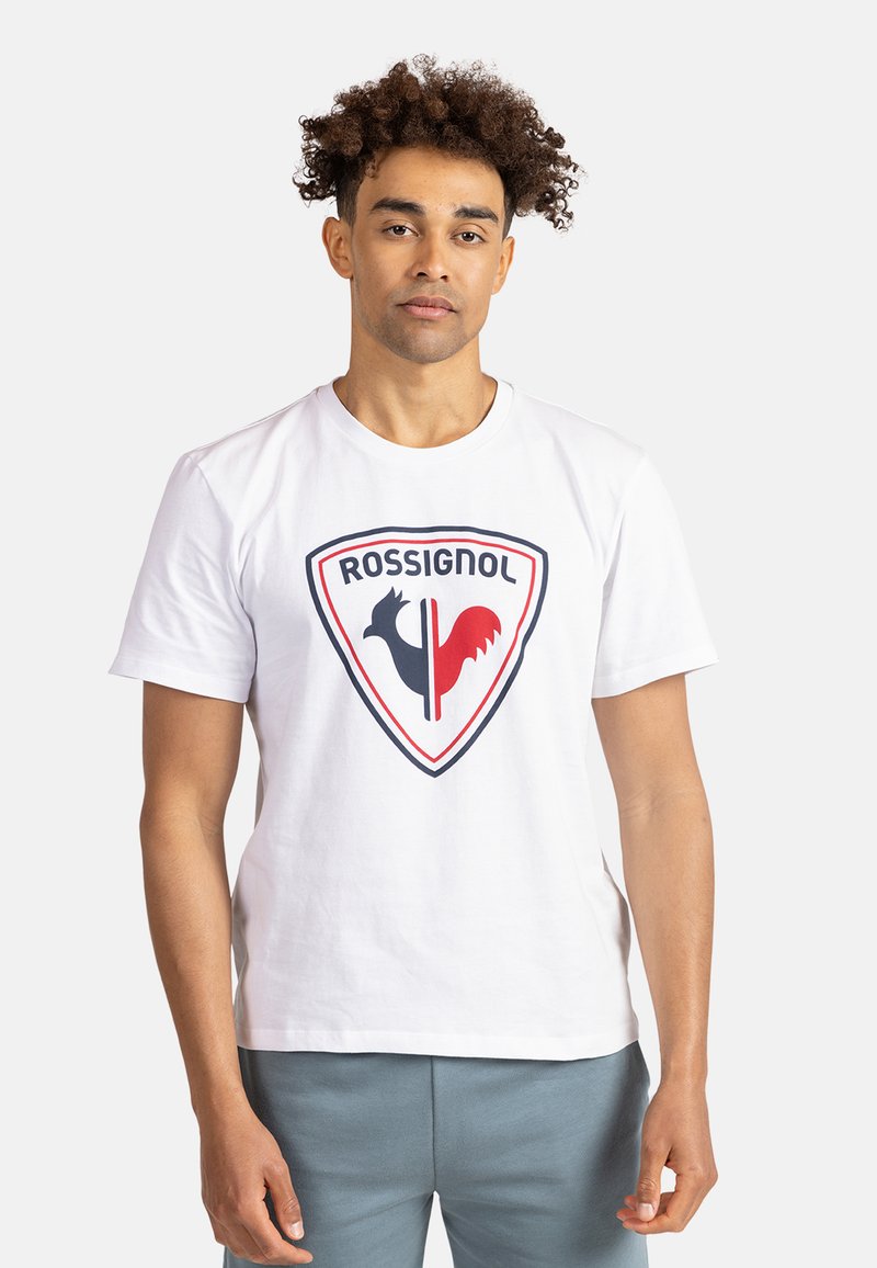 Camiseta de algodón blanca con un gran logo de escudo que presenta siluetas de gallos en negro y rojo y el texto "ROSSIGNOL" arriba. Diseño sencillo de cuello redondo.