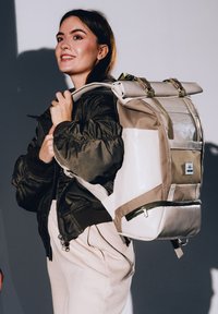 Világos bézs és szürke hátizsák, feltekerhető záródással, több zsebbel és zöld pántokkal. Tartós anyagból készült, sima tapintású.