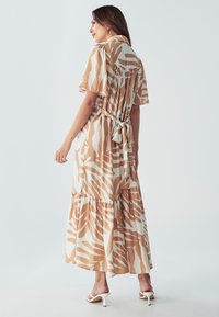 Hnedo-biely vzorovaný maxi šaty s krátkymi rukávmi, detailom na viazanie na chrbte a tiered lemom. Vyrobené z ľahkej látky. Viditeľné biele podpätky.