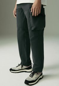 Pantaloni cargo gri închis cu o croială relaxată, având multiple buzunare și un material neted, ușor, asortați cu încălțăminte sport gri și neagră.