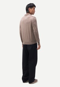 Junger Mann steht seitlich, trägt ein beiges Langarm-Poloshirt, schwarze weite Hose und taupefarbene Schuhe vor weißem Hintergrund.