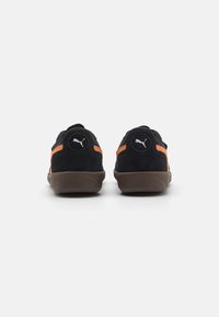 Les baskets en daim noir présentent des accents orange, un logo rond sur le talon et une semelle en caoutchouc marron. Design à dos fermé avec une texture lisse.