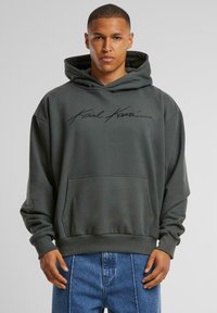 Sudadera gris oscuro con un bolsillo frontal y puños acanalados. Presenta una firma en negro en el pecho. Fabricada con tela suave y con un ajuste relajado.