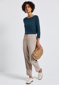 Ciemnoniebieska bluzka z długim rękawem, beżowe dopasowane spodnie z elastycznym paskiem, trzymająca dużą plecioną torebkę w kolorze brązowym, białe sneakersy z brązowymi akcentami.