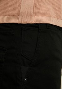 Černé kalhoty s texturovaným povrchem, vybavené přední kapsou a knoflíkovým detailem, spárované se světle růžovým žebrovaným svetr na úrovni pasu.