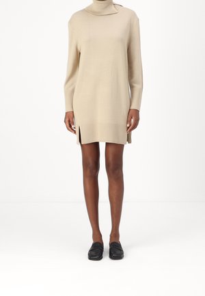 Kvinde iført en beige ribstrikket rullekravekjole med sidelommer og sorte loafers, stående mod en ensfarvet hvid baggrund.