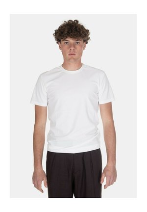 Giovane uomo con capelli ricci che indossa una semplice t-shirt bianca a maniche corte e pantaloni neri, in piedi davanti a uno sfondo bianco.