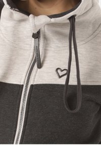 Sudadera con capucha gris y gris claro con cremallera, pequeño detalle de corazón, tejido bicolor y cordones, presenta una textura suave y un diseño deportivo.