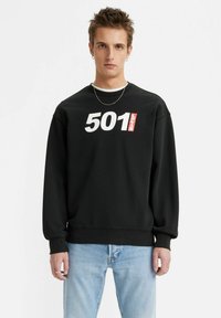 Zwarte sweatshirt met het "501" en "Levi's" logo in wit en rood, geribbelde manchetten en onderrand, ontspannen pasvorm, gedragen met lichtblauwe jeans.
