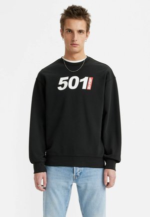 Czarna bluza z kapturem z białym i czerwonym logo "501" oraz "Levi's", ściągacze na rękawach i u dołu, luźny krój, noszona z jasnoniebieskimi dżinsami.